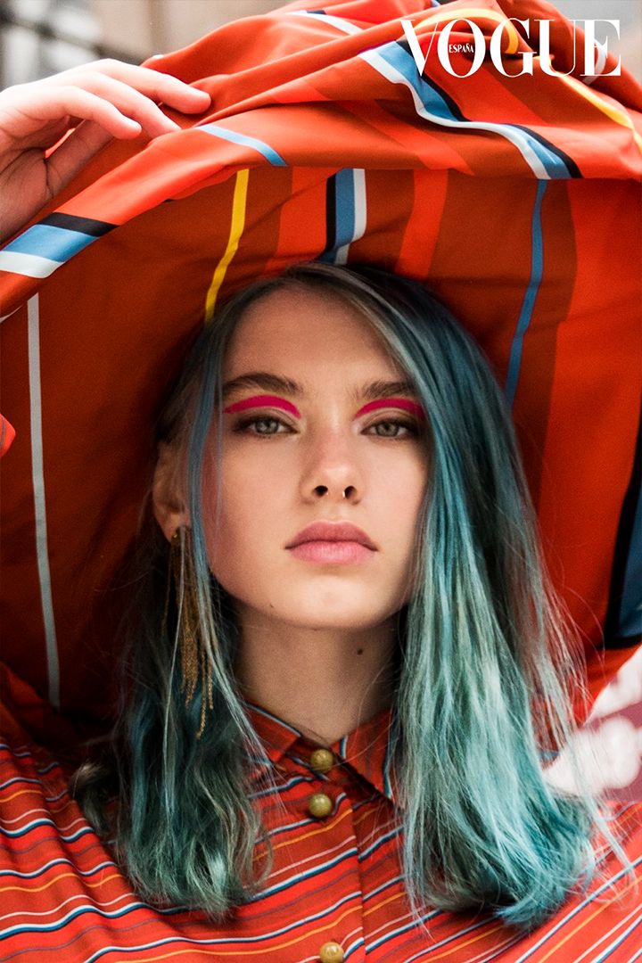 Close-up portrait of a young woman with blue-green hair and pink eye makeup, posing with red striped fabric and Vogue magazine logo.