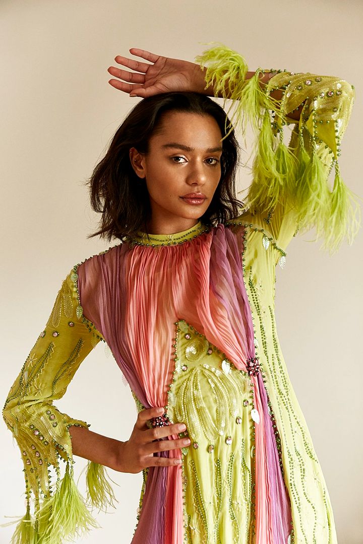 A young woman poses indoors wearing a light-green beaded gown with feather accents and a draped ombre scarf against a neutral backdrop.