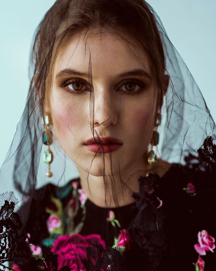 Close-up portrait of a woman with a black veil, wearing a floral top and statement earrings, set against a plain background.