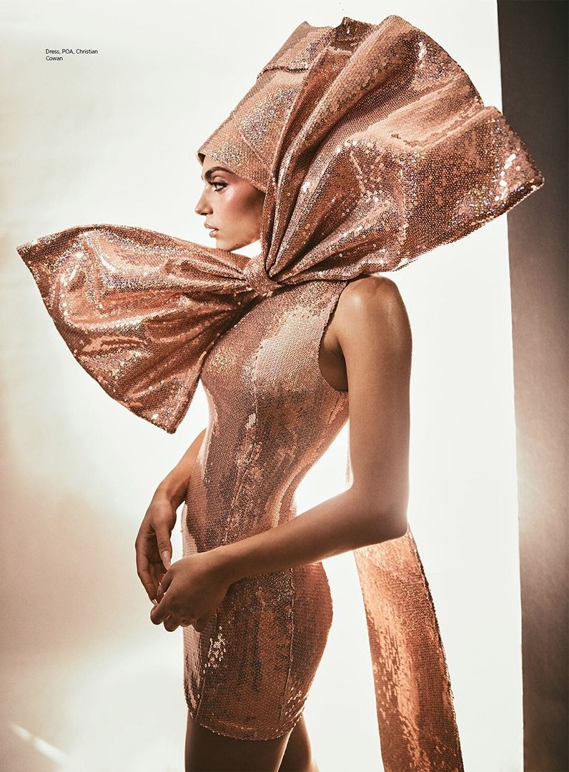Woman in a sequin dress with a large sequin bow on her head and shoulder stands in front of a brightly lit white background.