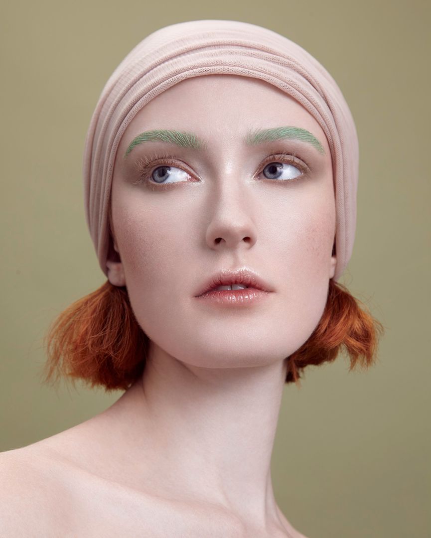 Headshot of a young woman with red hair and a beige headscarf against a muted green background. She has green eyebrows and is looking up and to the side.