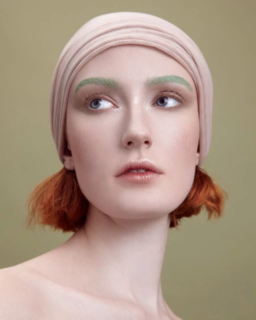 Headshot of a young woman with red hair and a beige headscarf against a muted green background. She has green eyebrows and is looking up and to the side.