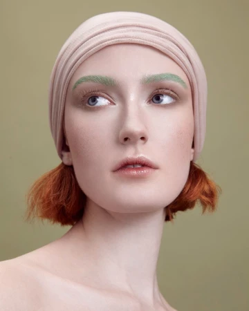 Headshot of a young woman with red hair and a beige headscarf against a muted green background. She has green eyebrows and is looking up and to the side.