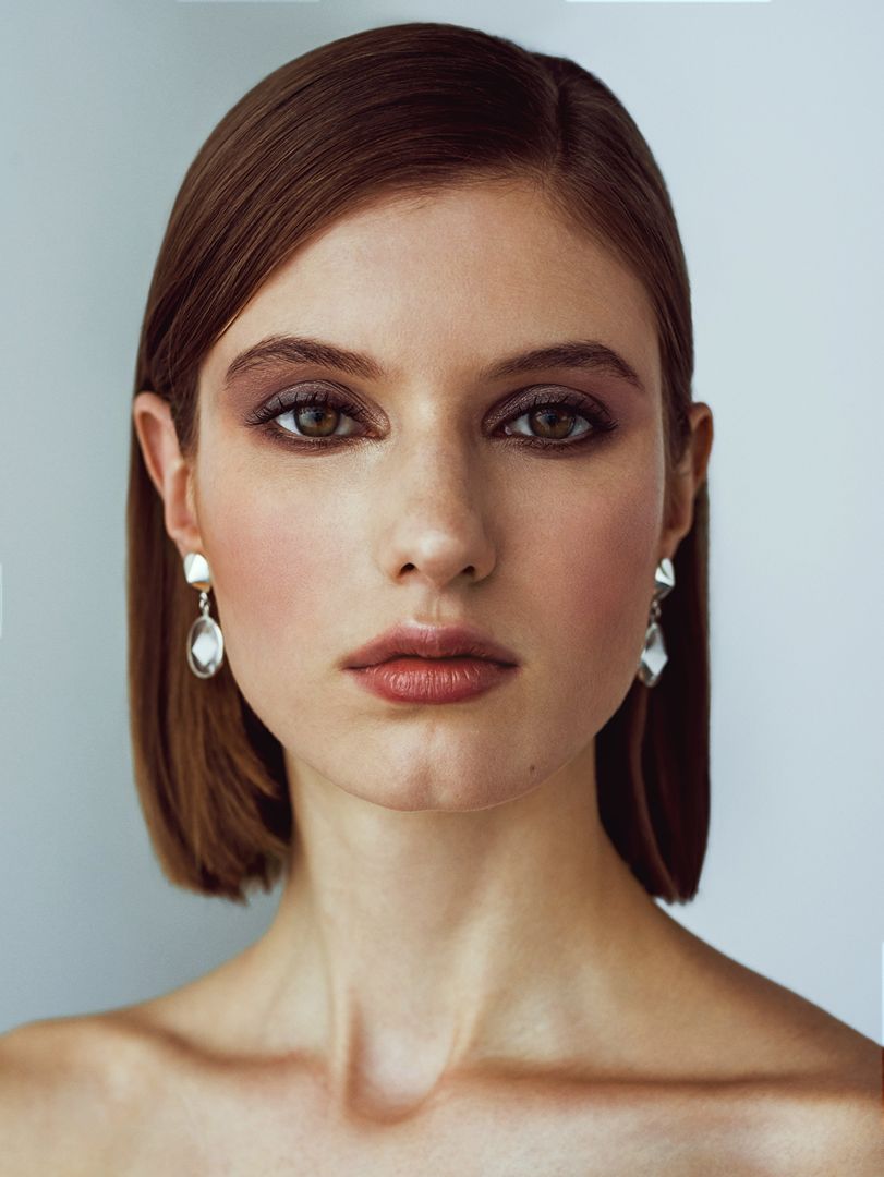 Close-up portrait of a woman with auburn hair and neutral makeup, wearing drop earrings against a plain grey background.