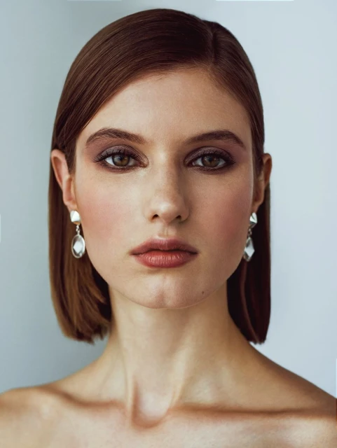 Close-up portrait of a woman with auburn hair and neutral makeup, wearing drop earrings against a plain grey background.