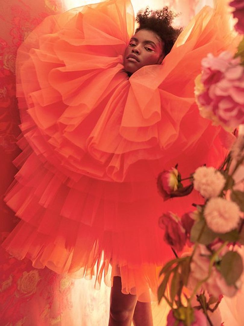 Woman in a large, layered orange gown poses in front of a floral backdrop with pink and white flowers in a fashion editorial shot.