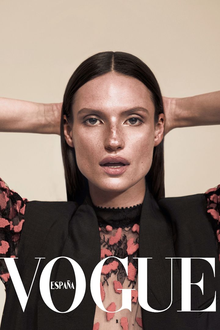 Portrait of a woman with dark hair wearing a black vest over a floral shirt, posing against a tan background, Vogue España logo visible.