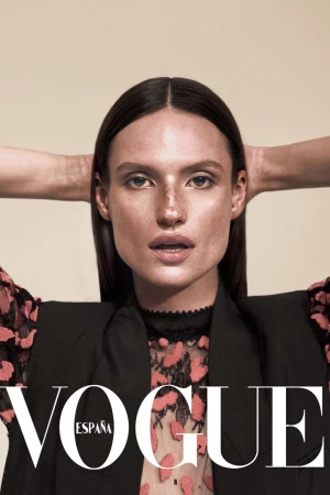 Portrait of a woman with dark hair wearing a black vest over a floral shirt, posing against a tan background, Vogue España logo visible.