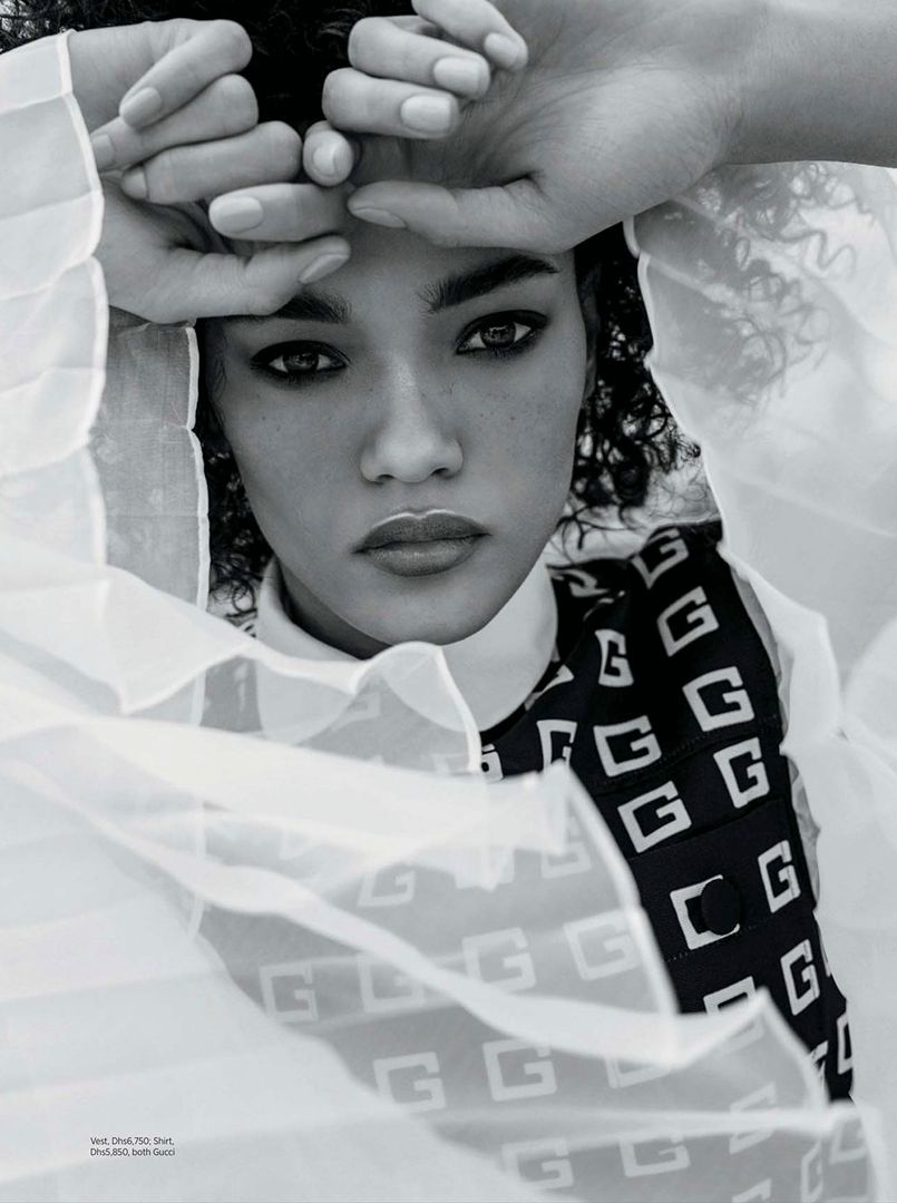 Close-up portrait of a woman with dark curly hair holding her hands to her forehead, framed by sheer white fabric.