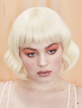 Close-up studio shot of a fair-skinned young woman with blonde bob hairstyle and coral makeup against a peach backdrop, portrait orientation.