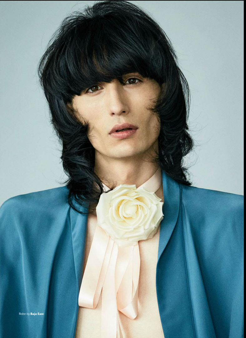 Portrait of a young adult male with dark hair wearing a blue jacket and peach-colored shirt with a cream rose pin. Fashion shoot.