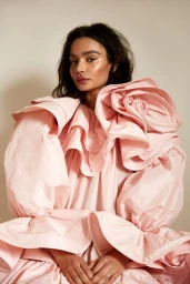 Model with dark hair wearing a voluminous pink ruffled dress against a plain studio background