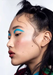 Close-up profile portrait of a woman with vibrant blue eyeshadow, rosy cheeks, and dark hair styled in small buns.
