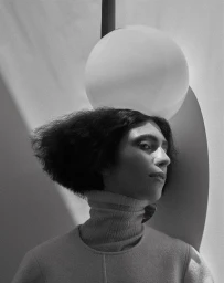 Monochrome portrait of a woman in a turtleneck posing with a large spherical globe light behind her head.