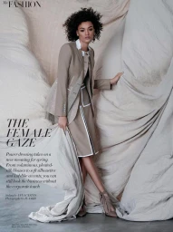 Model with dark curly hair wearing a taupe suit jacket and matching skirt against a draped fabric background.
