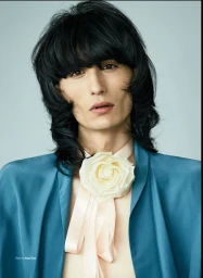 Portrait of a man with dark shaggy hair wearing a teal jacket and a large white rose choker necklace.