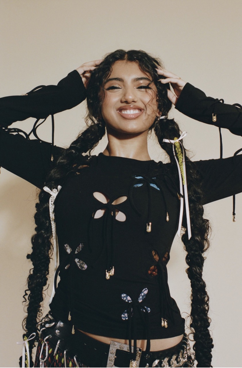 Smiling woman poses with hands in hair in a studio shot, wearing a black cut-out top with butterfly accents and accessorized pigtails.
