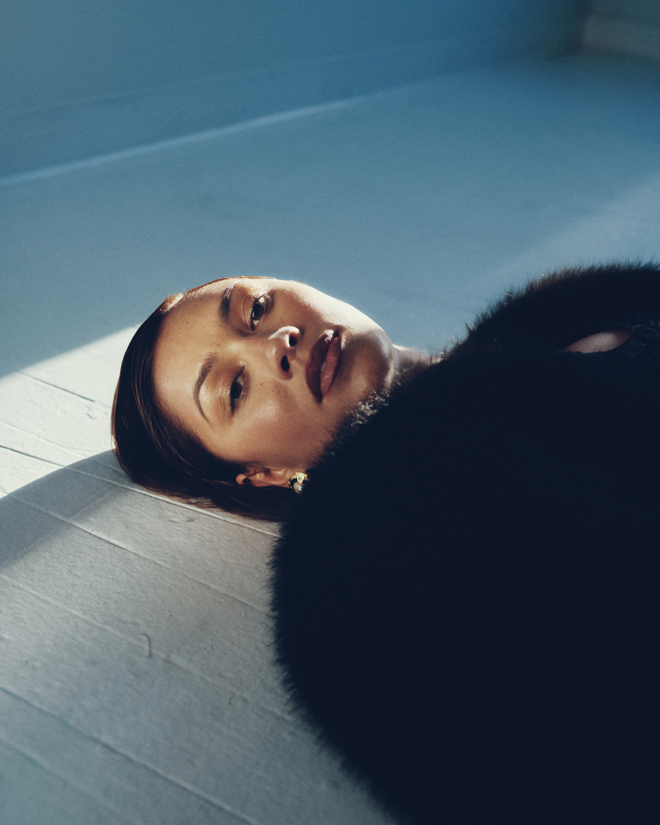 Elegant woman lies on a hardwood floor wearing a fur coat. Soft lighting accentuates her face in a studio-like environment.