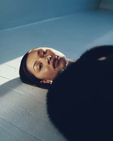 Elegant woman lies on a hardwood floor wearing a fur coat. Soft lighting accentuates her face in a studio-like environment.