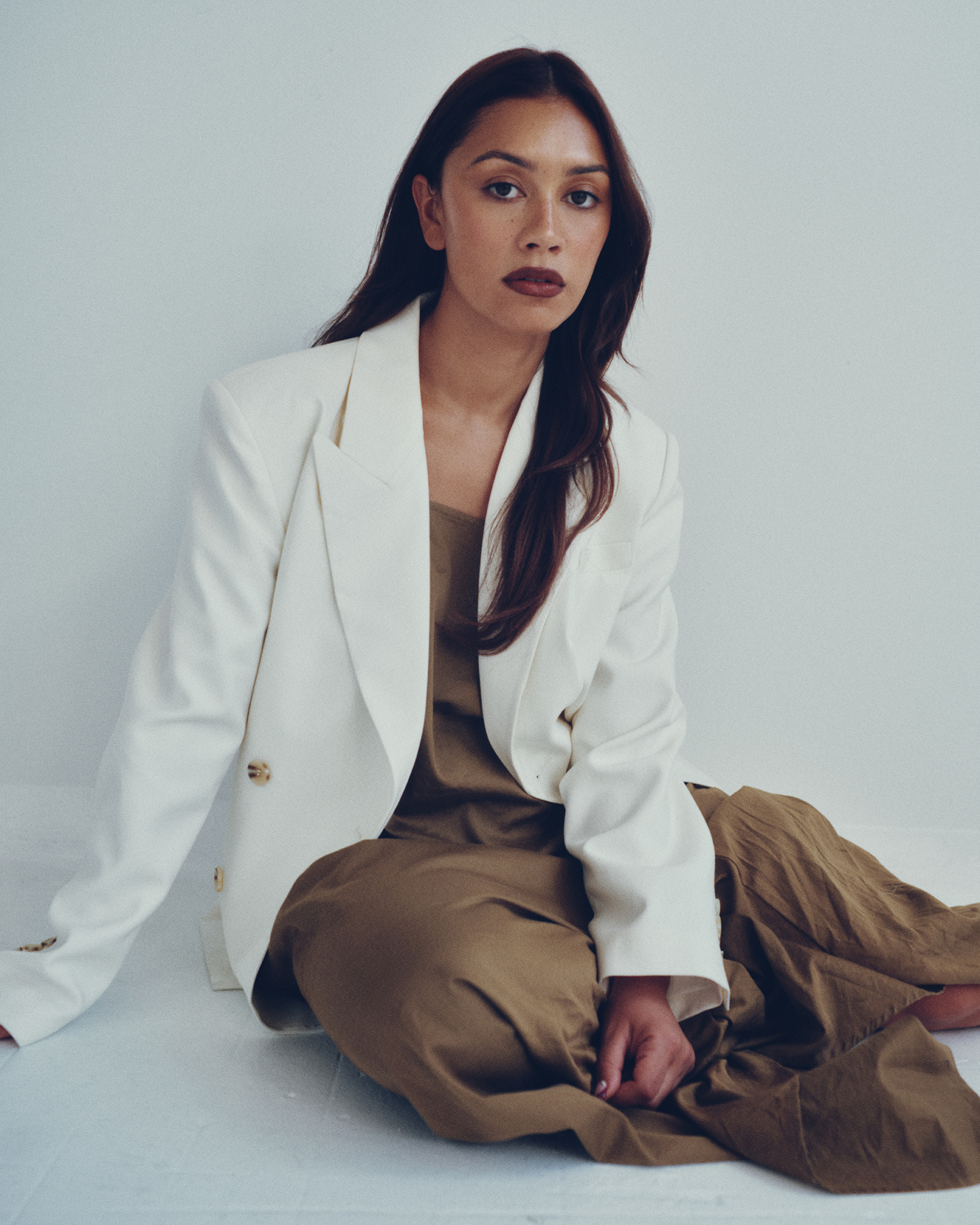 Portrait of an adult female in a beige dress and white blazer sitting against a white backdrop in a studio environment.
