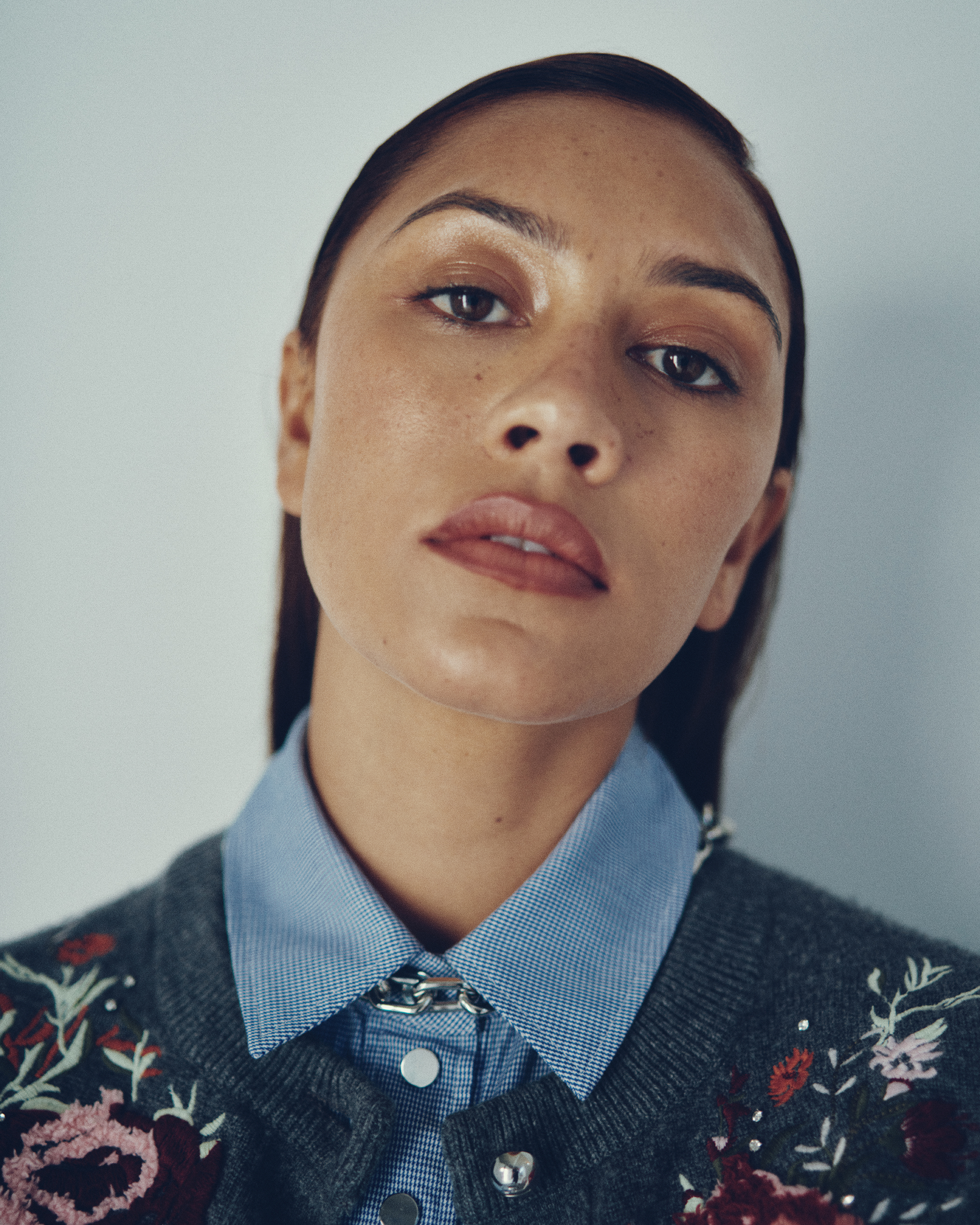 Portrait of a woman with fair skin and long dark hair, wearing a floral cardigan and collared shirt, against a light grey background.