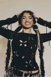 Portrait of a smiling woman with long braided pigtails wearing a black cut-out top styled with ribbons and charms.
