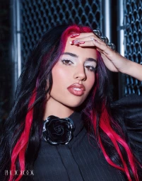 Close-up portrait of a woman with dark hair featuring vivid pink streaks, wearing a black collared top with a rose brooch against a chain-link fence