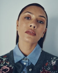 Close-up portrait of a woman wearing a blue buttoned collar shirt and embroidered grey cardigan against a light background.