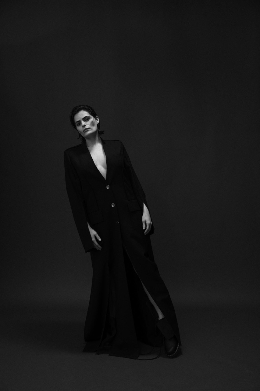 A woman in a black jacket and long skirt poses in a studio with dark background. Full shot in black and white, fashion editorial.