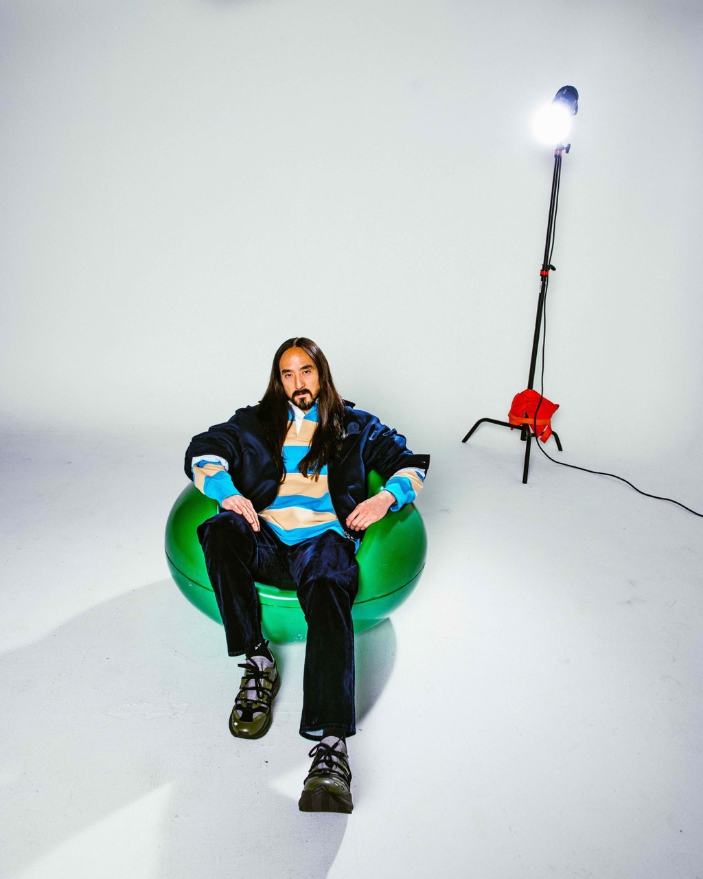 Adult male with long dark hair sits in a green inflatable chair, wearing a striped top and dark pants in a photo studio with lighting equipment.