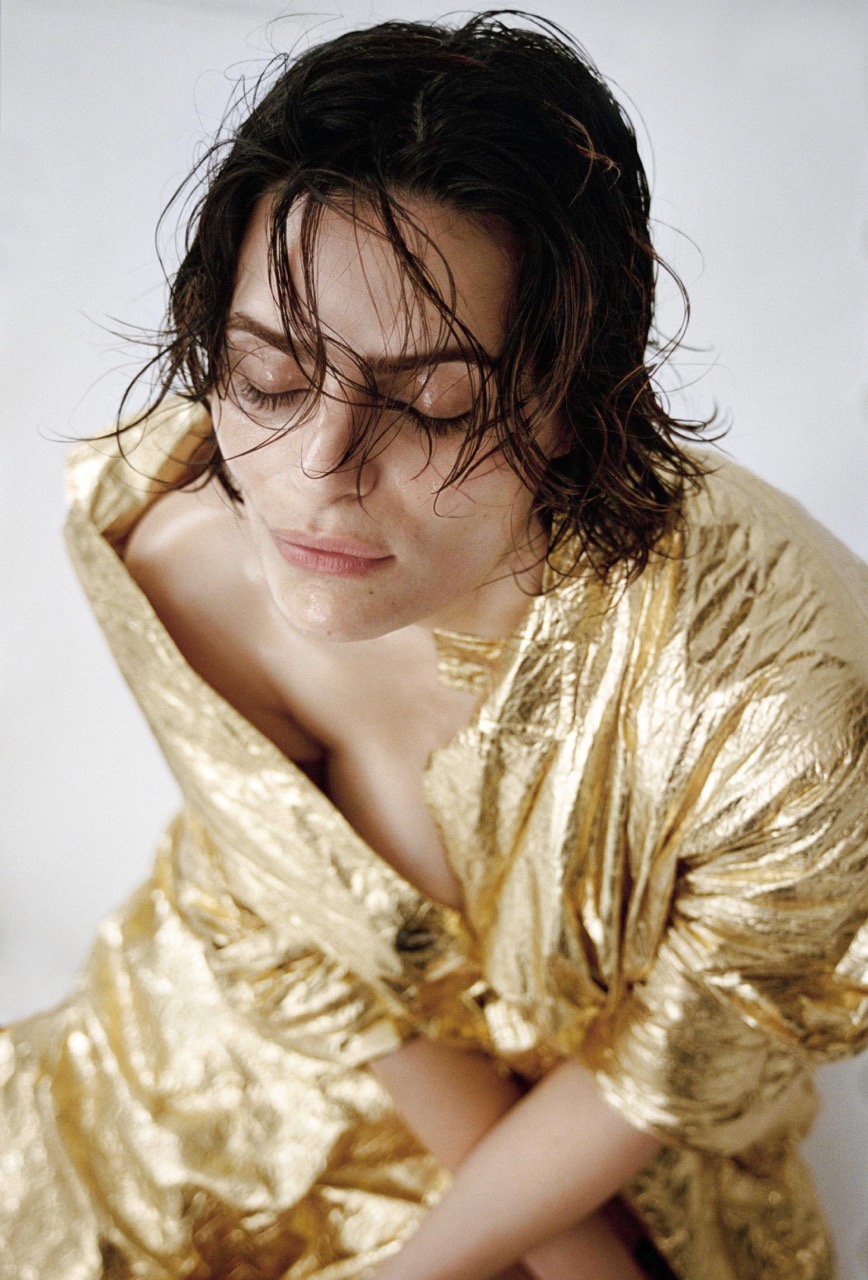 Portrait of a brunette adult female with closed eyes, draped in a shimmering gold textile against a neutral background in a studio setting.