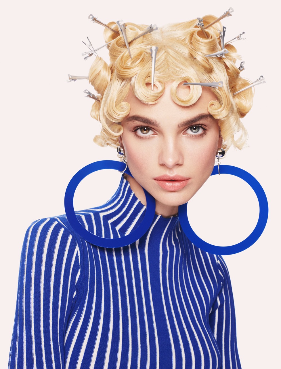 Portrait of a woman with blonde hair styled in rollers, wearing large blue hoop earrings and a blue and white striped top.