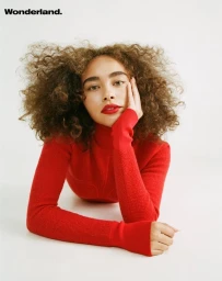 A woman with curly brown hair wearing a vibrant red turtleneck sweater poses against a plain white background.