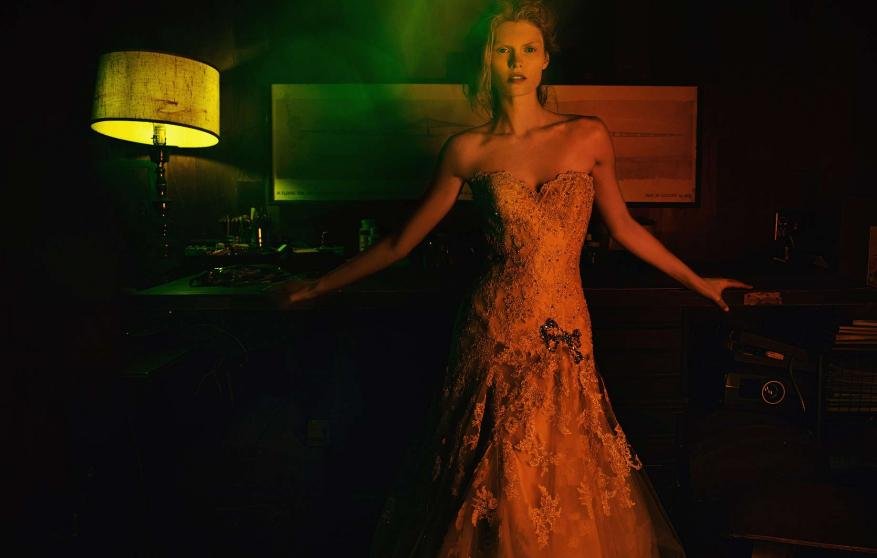 Adult female fashion model in a strapless orange lace gown with bow in dark indoor setting with floor lamp and green and orange lighting.