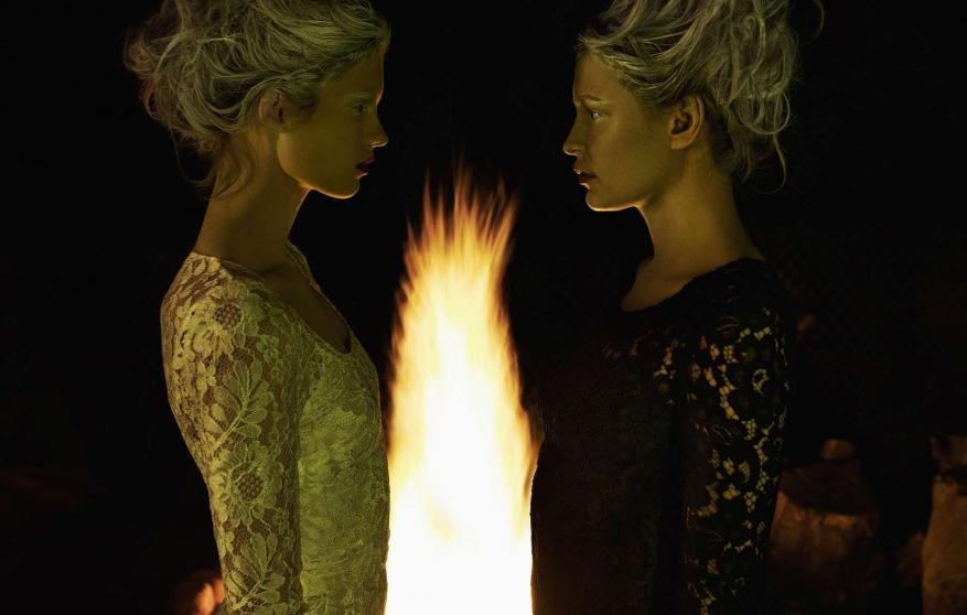 Two fair-skinned young women face each other with a bright flame between them against a dark background; warm and cool tones.