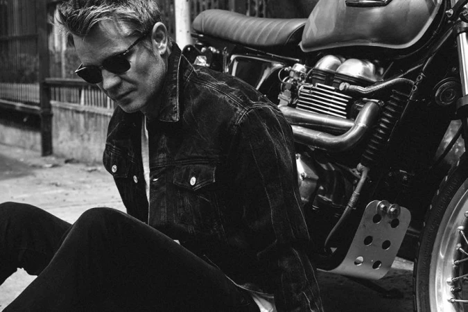 Man wearing sunglasses and a denim jacket seated next to a vintage motorcycle in a black and white photograph