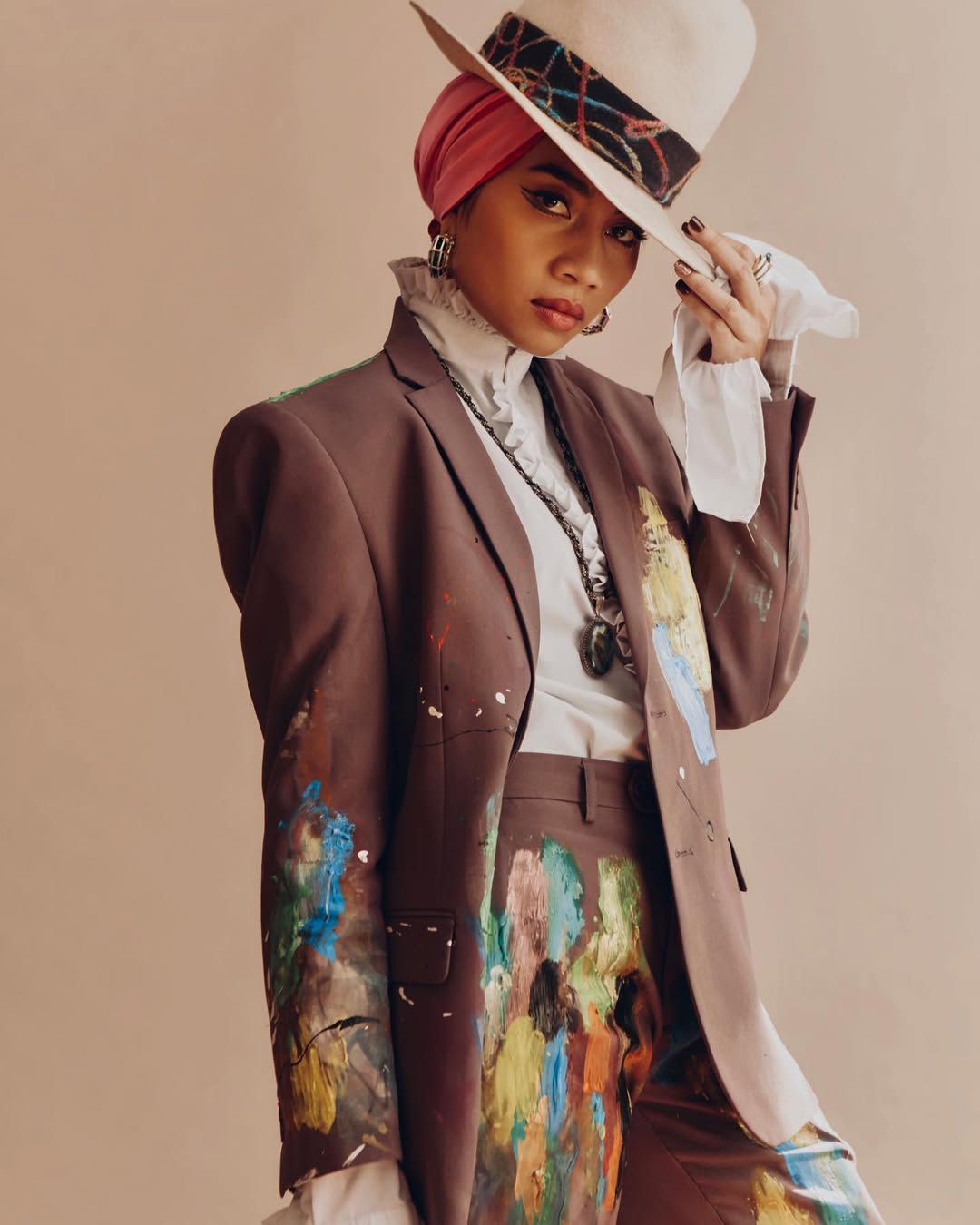 A woman in a brown suit and hat poses with paint splatters on her clothing, standing against a neutral background in a stylish studio shot.