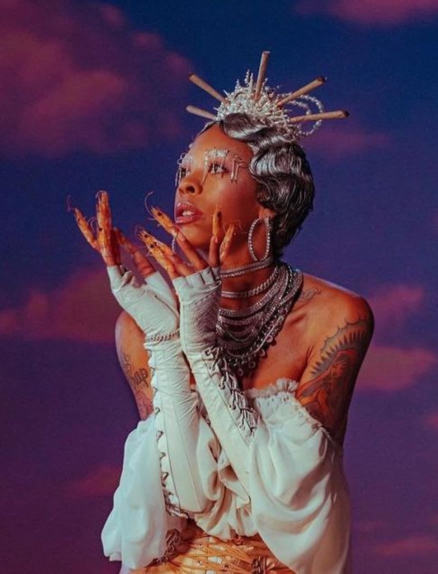 Portrait of a woman in an ornate crown with long fingernails, multiple necklaces, and white top in front of a sky background.