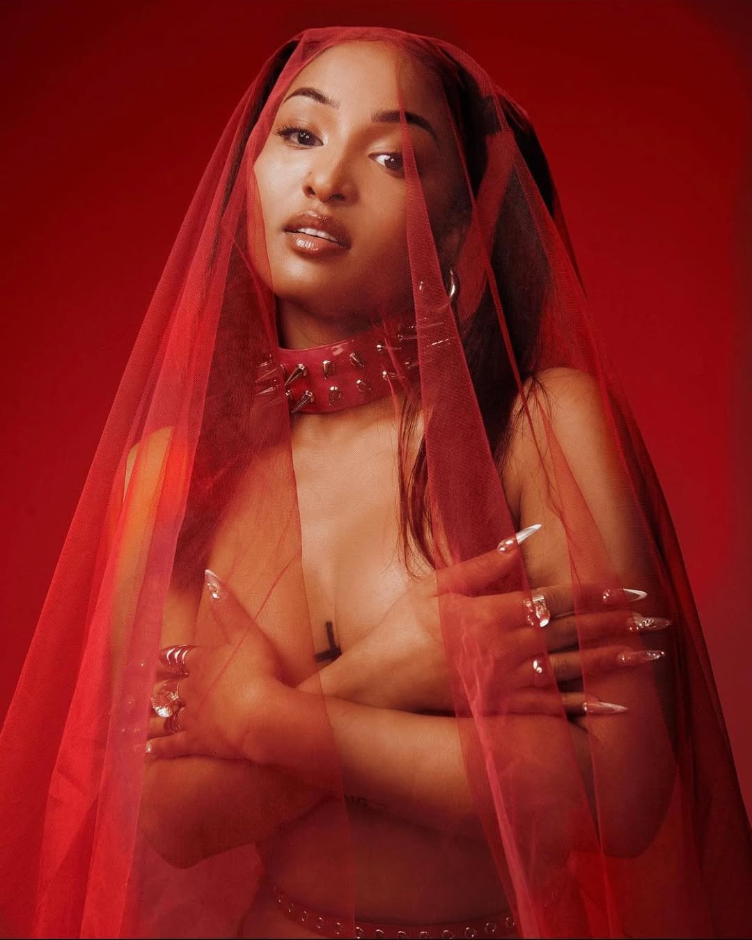 Portrait of a woman with dramatic eye makeup, wearing a red veil and spiked choker, against a deep red background in studio lighting.