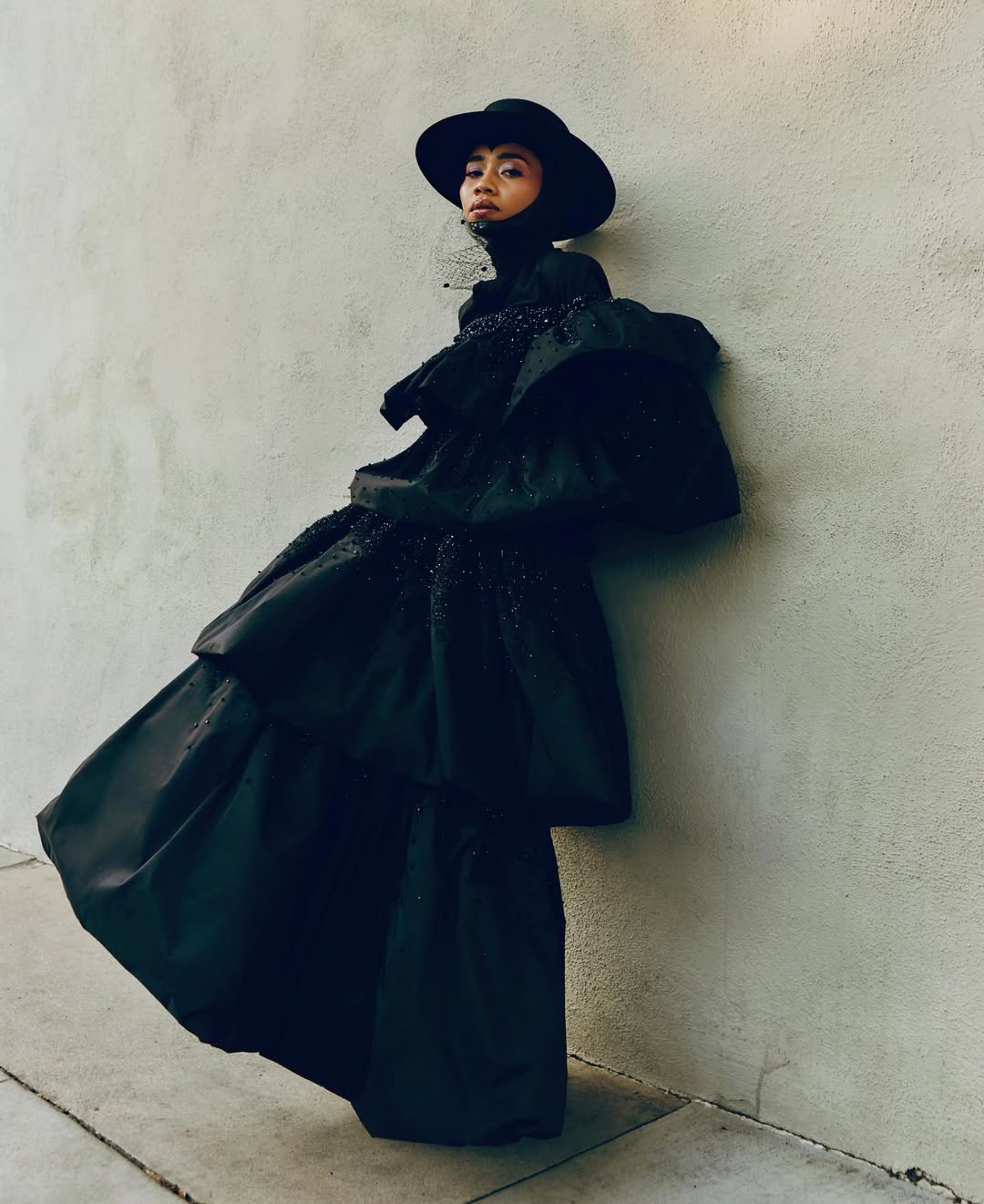 A woman in a large black dress and hat leans against a wall. The attire is extravagant, the setting minimal, and the lighting is soft.