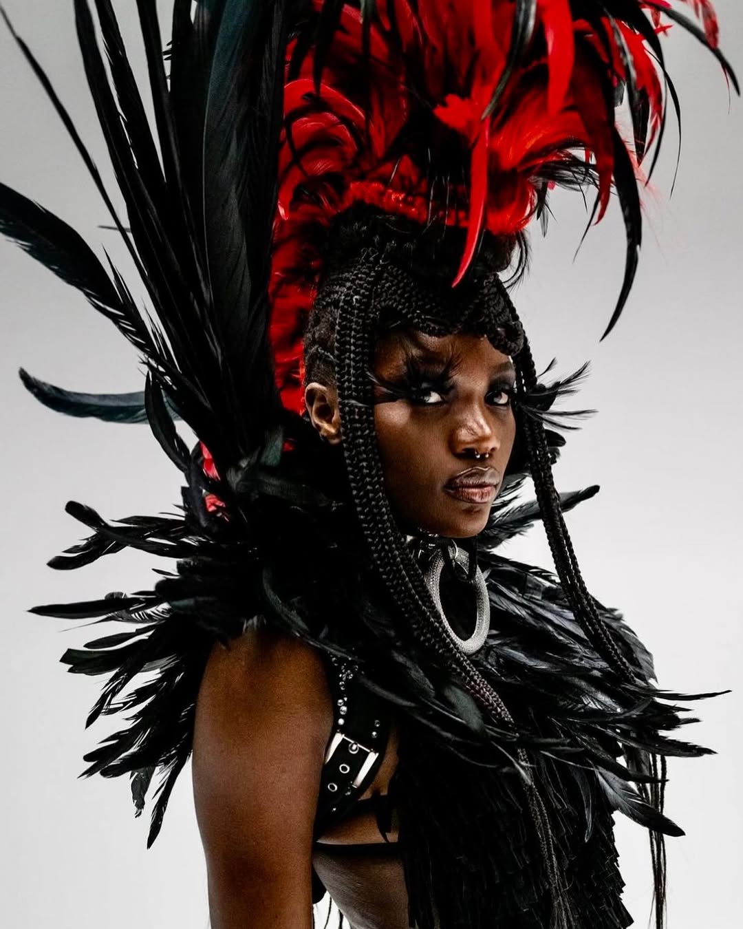Portrait of an adult female with dark skin wearing a feathered headpiece and collar in black and red against a white background.