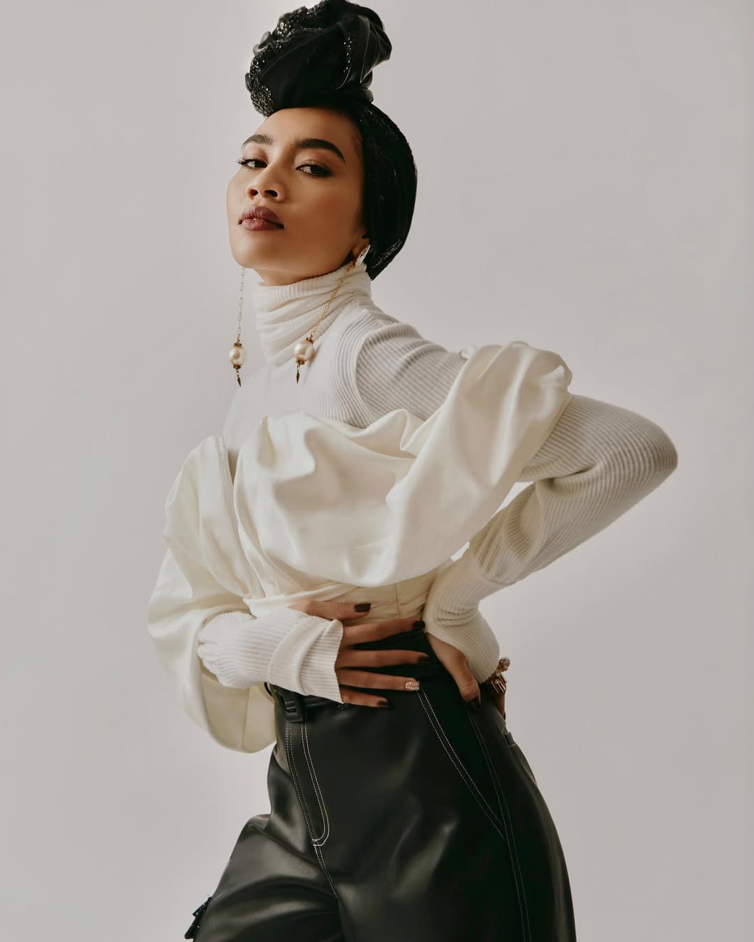 A woman in a fashionable white blouse and black pants poses in a studio with a neutral backdrop, her hand on her hip.
