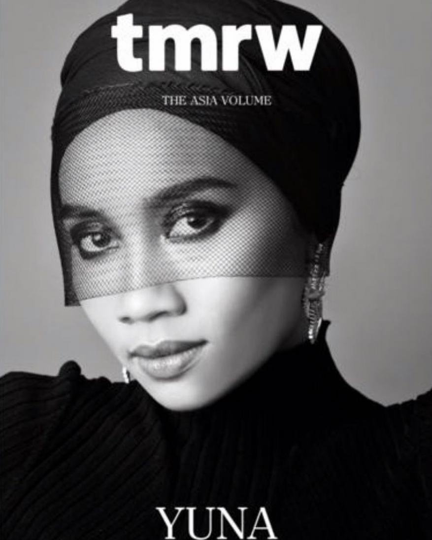 Black and white portrait of a person with dark hair, covered with a black headscarf and veil, against a light gray background.