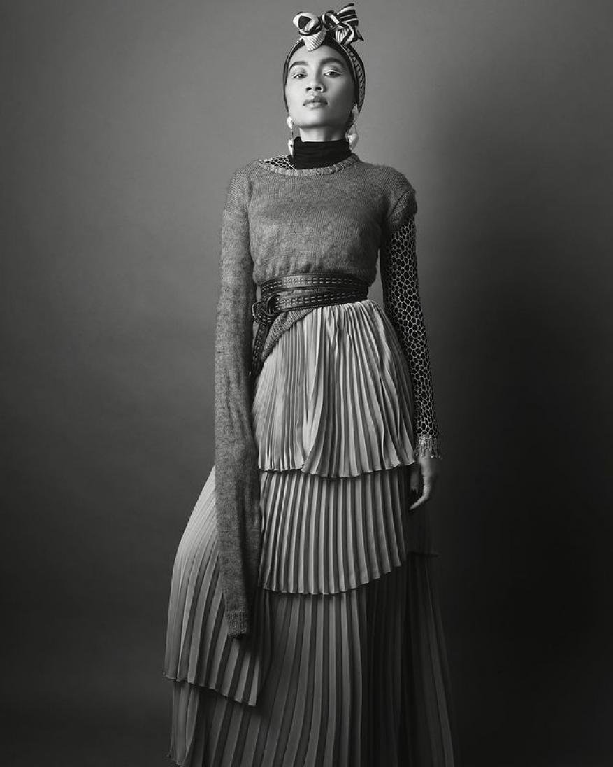 Monochrome portrait of a stylish woman wearing a headscarf, sheer sweater, layered pleated skirt, and statement earrings against a gray background.