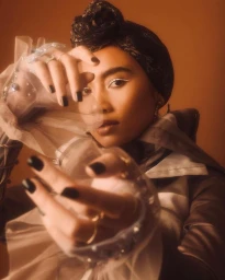 Model wearing a dark headwrap and sheer ruffled blouse with dark fingernails posing with hands raised in a warm-toned portrait