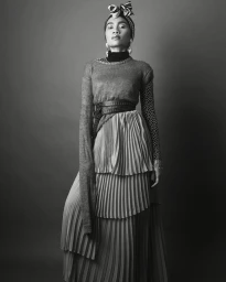 Monochrome fashion portrait of a woman wearing a patterned headwrap, layered pleated skirt, and double belt styled by Aquiles.