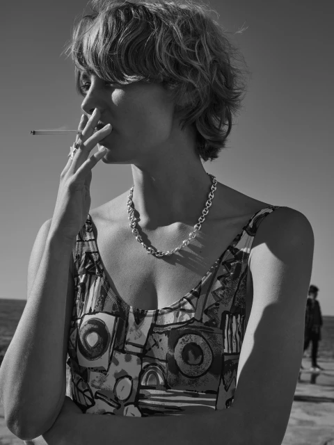 Monochrome portrait of a young adult woman smoking a cigarette outdoors, wearing a patterned top and necklace against a clear sky.