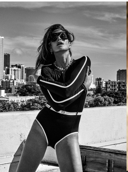 Monochrome outdoor portrait of a woman wearing a retro-style swimsuit, sunglasses, and jewelry, posing with a city skyline backdrop.