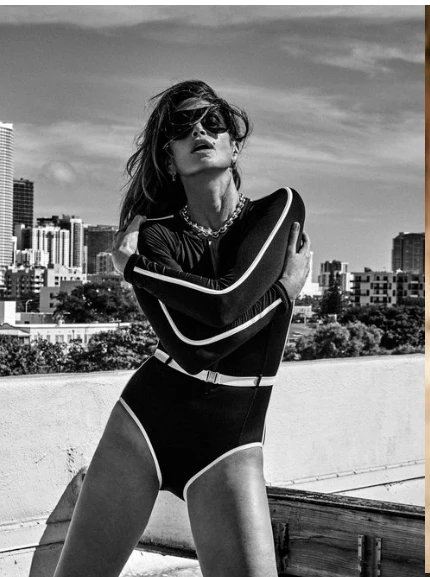 Monochrome outdoor portrait of a woman wearing a retro-style swimsuit, sunglasses, and jewelry, posing with a city skyline backdrop.