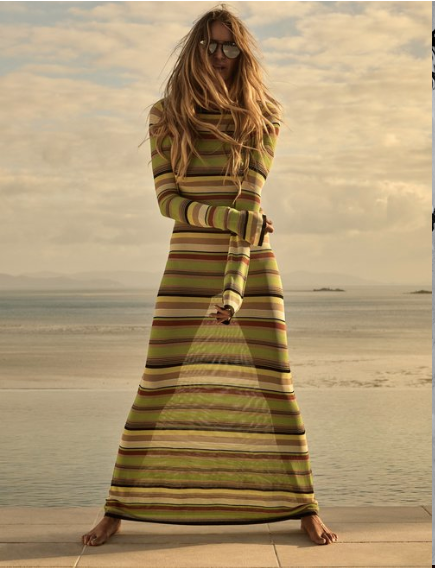 A tall, fair-skinned woman wearing sunglasses and a striped maxi dress poses barefoot on a sunlit stone patio with a sea view.